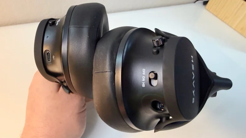 Hand holding large black over ear headphones with thick cushioned ear cups, USB C charging port, power switch labeled Off On HB, and a protruding attachment on one side