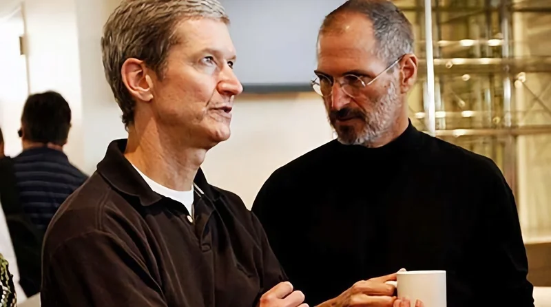 Two middleaged men stand close together indoors, conversing; one faces forward with arms crossed, the other in glasses and dark clothing holds a white mug, background slightly blurred