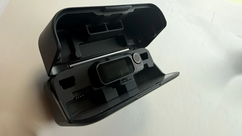 Open hard case holding a compact electronic device with a small screen and circular control knob, resting in a molded interior tray against a plain light background.