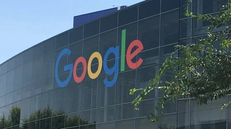 Large glass office building with the multicolored Google logo on its facade, set against a blue sky, partially framed by green tree branches in the foreground