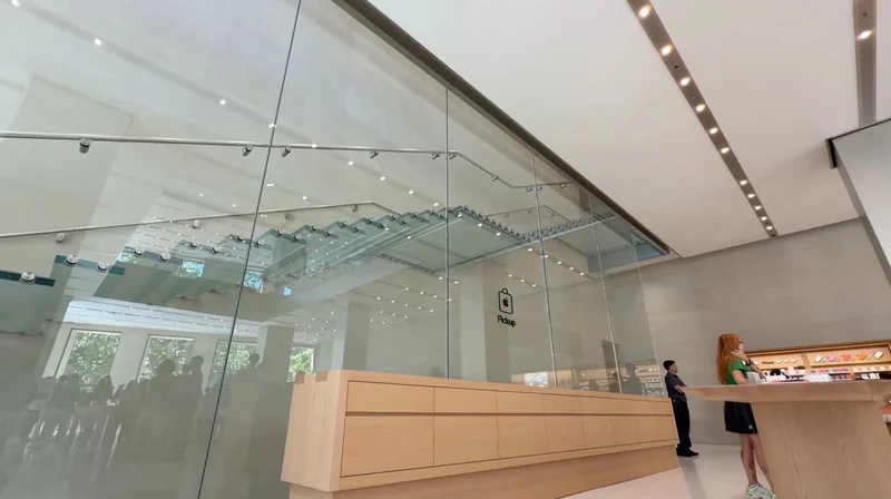 Spacious modern tech store interior with high glass walls, minimalist wooden counters, a glass staircase overhead, bright ceiling lights, and a few people standing near the counters