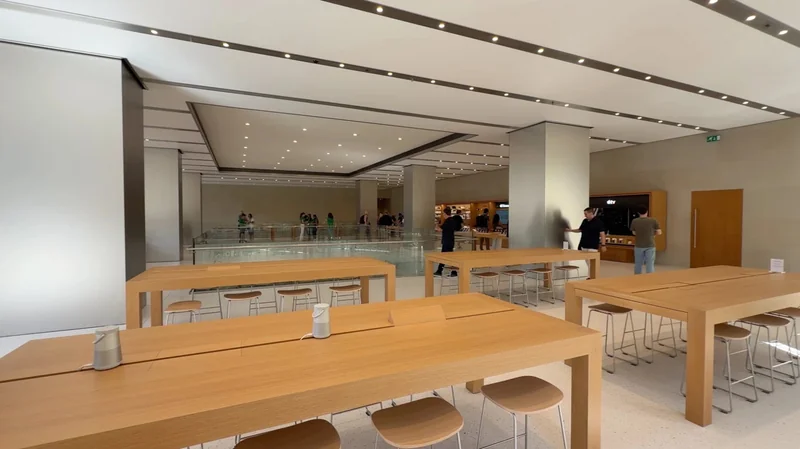 Spacious modern tech store interior with long light-wood tables, stools, bright ceiling lights, and a few people browsing in the background near product displays and a glass railing