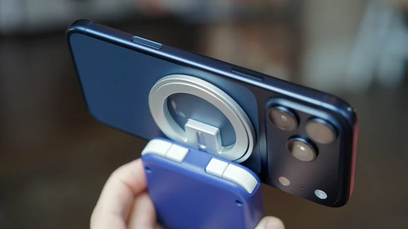 Hand holding a purple handheld gadget attached magnetically to the back of a dark smartphone with a circular metal stand, showing dual rear cameras in a blurred indoor setting