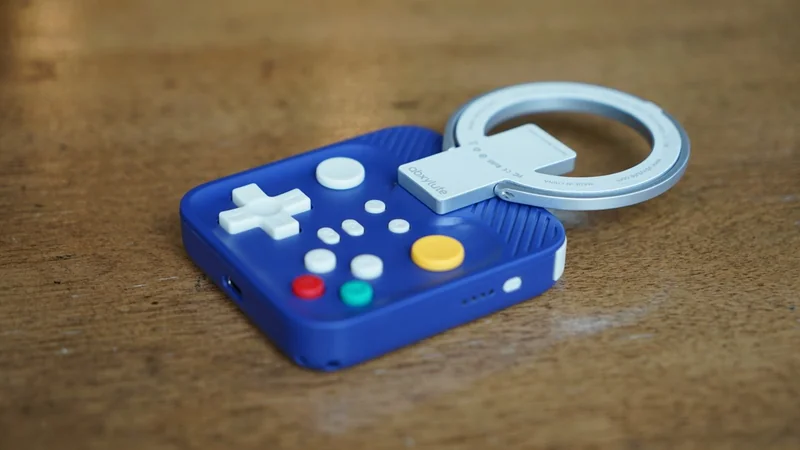 Small purple handheld game controller with colorful buttons and white directional pad, attached to a large circular silver keyring, resting on a wooden surface