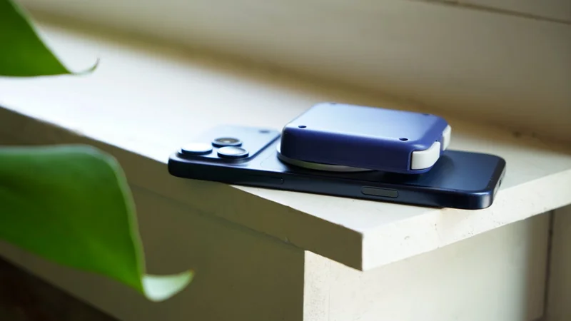 iPhone lying on a windowsill with a small rectangular purple controller resting on top, next to a green leafy plant in soft natural light