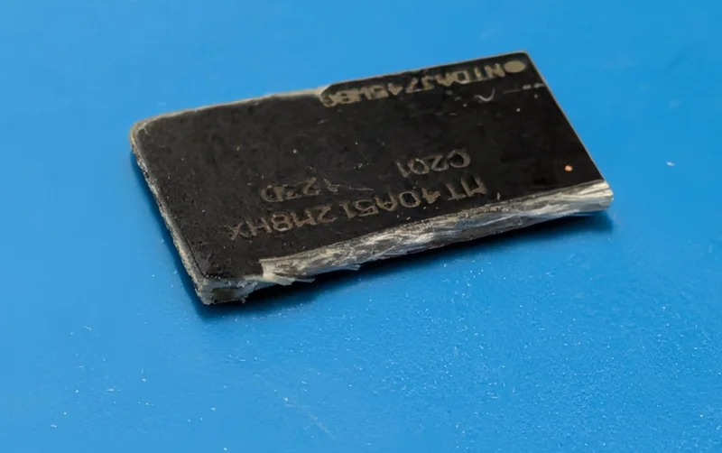 A broken semiconductor chip marked with 'NT04GA5128HX' lies on a blue surface.