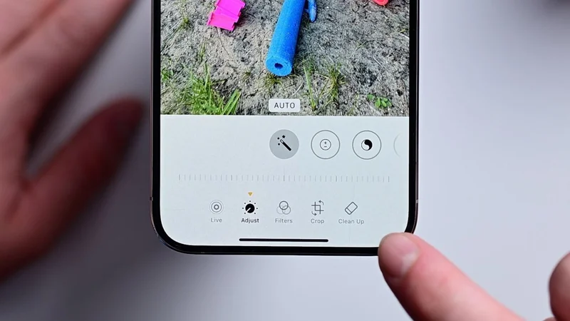 Close-up of a smartphone screen in a photo editing app, showing adjustment tools and sliders, with a finger pointing at the bottom controls and blurred hand in background