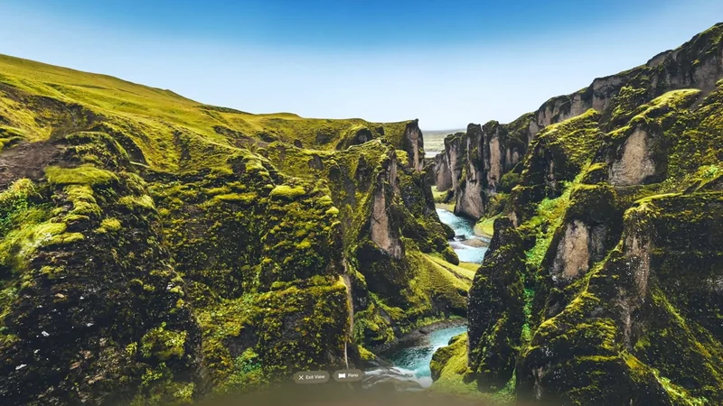 Lush green moss-covered cliffs form a deep canyon with a winding blue river below under a clear bright sky, suggesting a remote, dramatic natural landscape