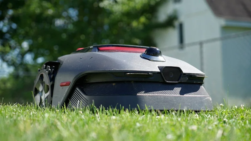 Low-angle view of a robotic lawn mower moving through green grass in a sunny backyard, with a house, fence, and leafy trees blurred in the background