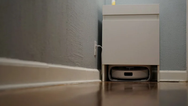 Low hallway view of a small white cabinet with a robot vacuum docked beneath it against gray walls and wooden floor, power cord plugged into wall outlet nearby