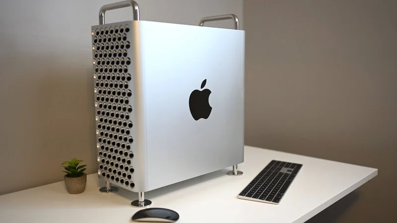 Silver Mac Pro desktop tower with perforated side and Apple logo on a white desk, beside a slim keyboard, wireless mouse, and small potted plant against a plain wall