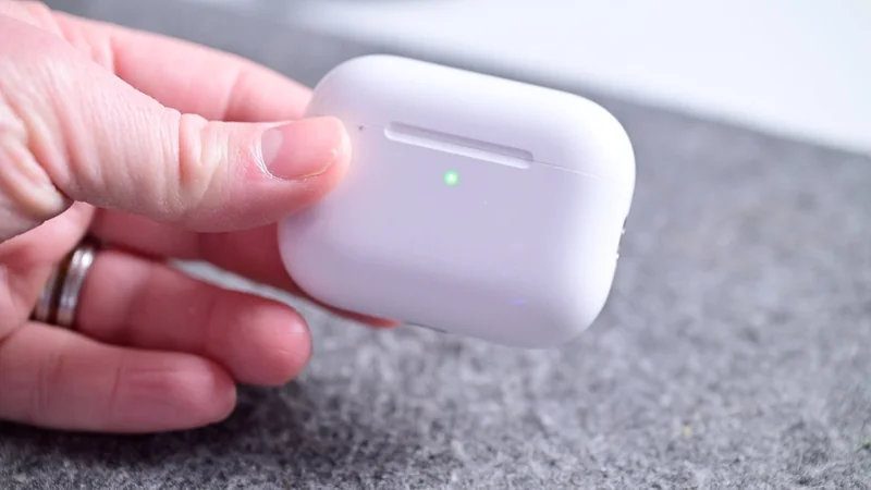 Hand holding AirPods Pro 3 wireless earbuds charging case on a gray surface, with a small green light glowing on the front of the case.