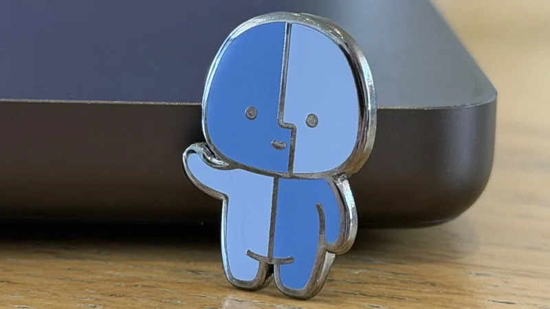 Small blue and light blue cartoon character pin with simple facial features, standing and waving on a wooden surface next to the corner of a dark electronic device