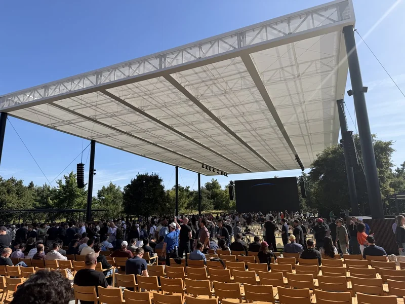 Outdoor event venue with a large white canopy, rows of empty yellow chairs in front, a crowd standing near the stage area, and trees and blue sky in the background