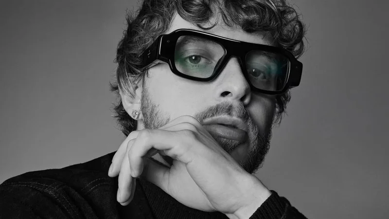 Young adult with curly hair and beard wearing large dark glasses, resting chin on hand, gazing thoughtfully toward the camera in a black-and-white close-up portrait
