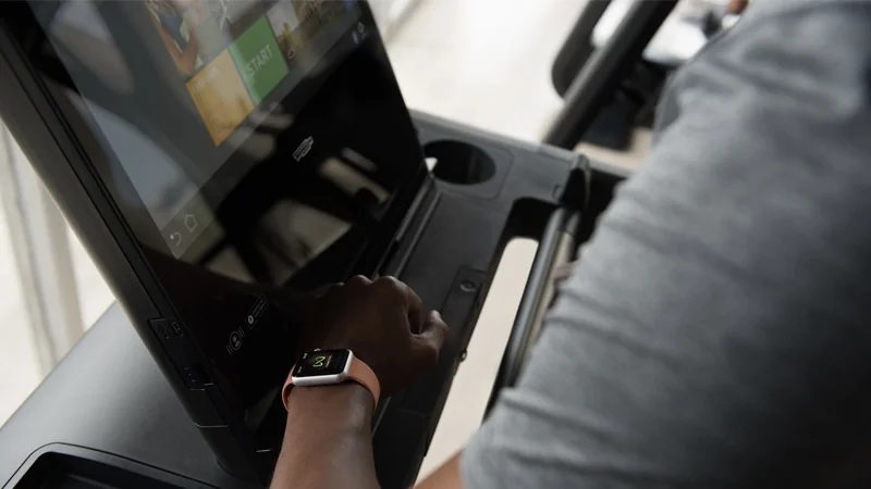 Person using a touchscreen fitness machine while wearing a smartwatch, pressing buttons on the console next to a large display and cup holder in a bright indoor setting