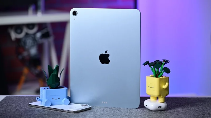 iPad Air M4 standing upright on a table, flanked by two small cartoon-style plant pots and resting on a smartphone, with a softly blurred purple and blue background