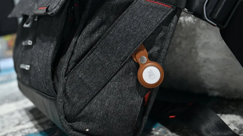 Dark gray backpack on the floor with an Apple AirTag 2 in a brown holder clipped near a side pocket, showing part of the shoulder strap and soft background cushion