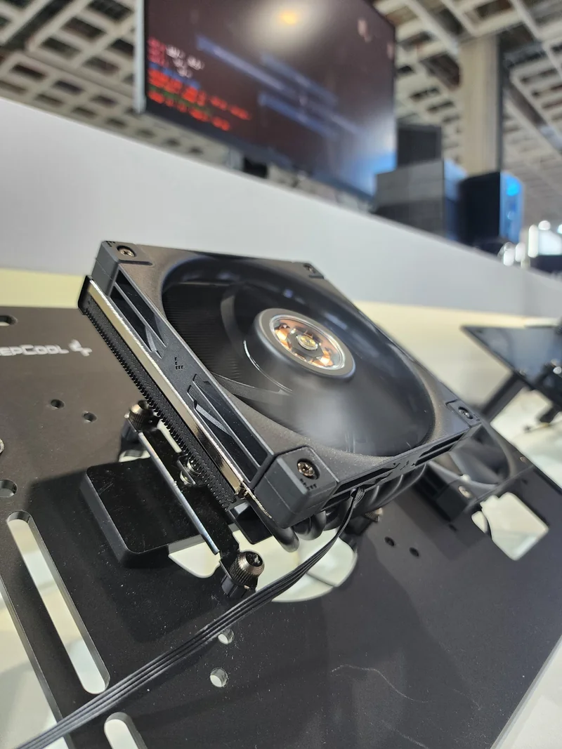 A DeepCool fan is mounted on a test bench with a blurred screen displaying lines of code in the background.