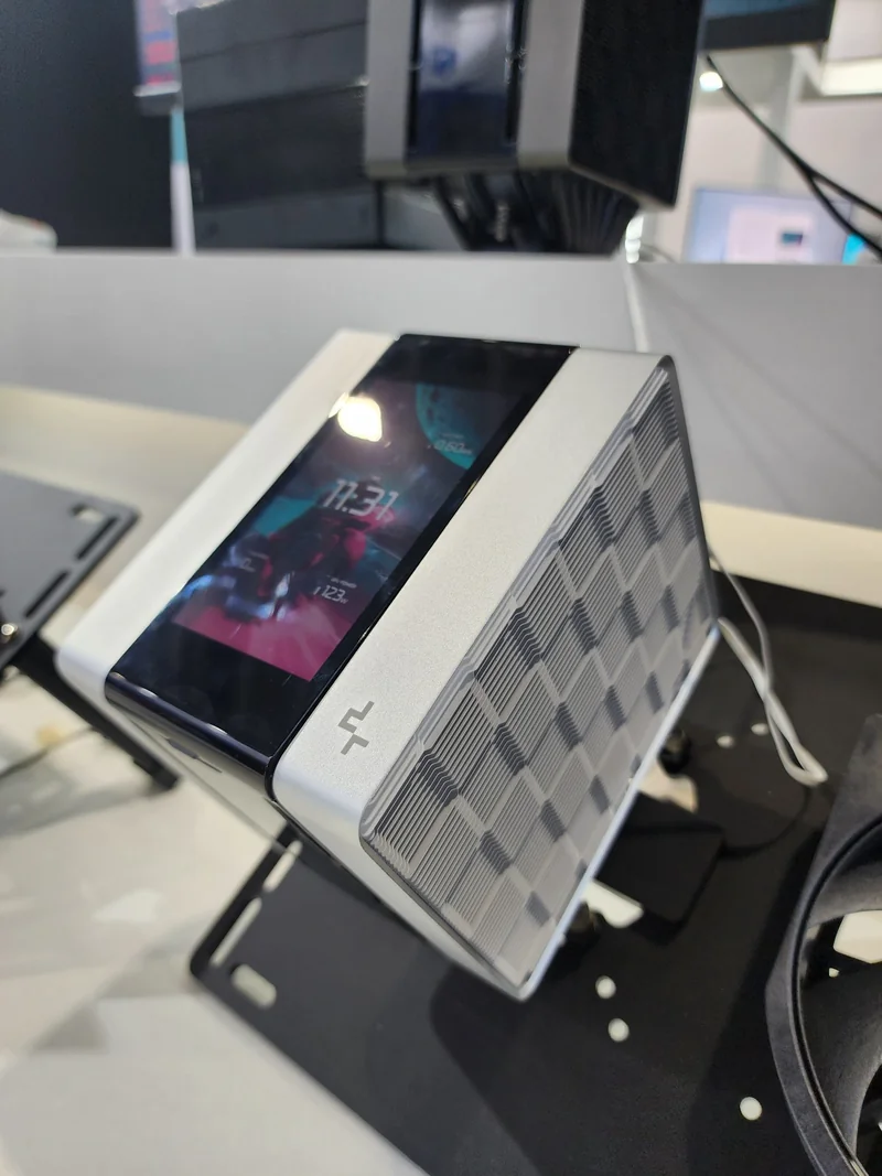 A compact PC with a built-in screen displaying '11:37' is housed in a sleek, silver chassis with a geometric pattern and a logo on the corner.