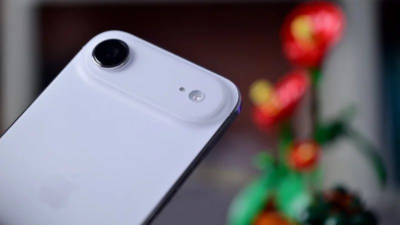 Close-up of a white smartphone's back camera and flash, with an Apple logo faintly visible and colorful, blurred flowers in the background