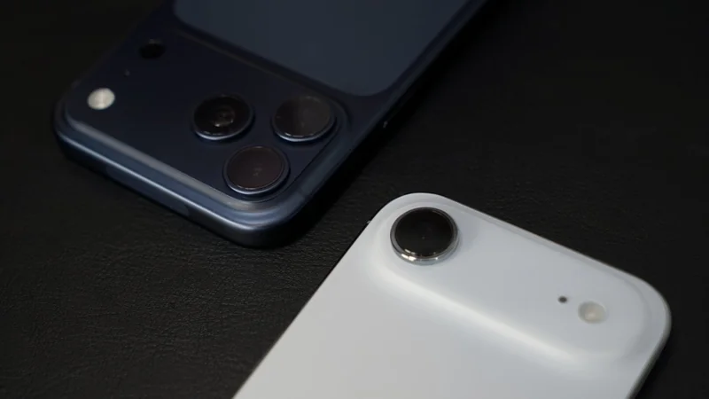 Two smartphones on a dark surface, one navy with three rear cameras and one white with a single raised camera lens, both shown from the back focusing on their camera modules