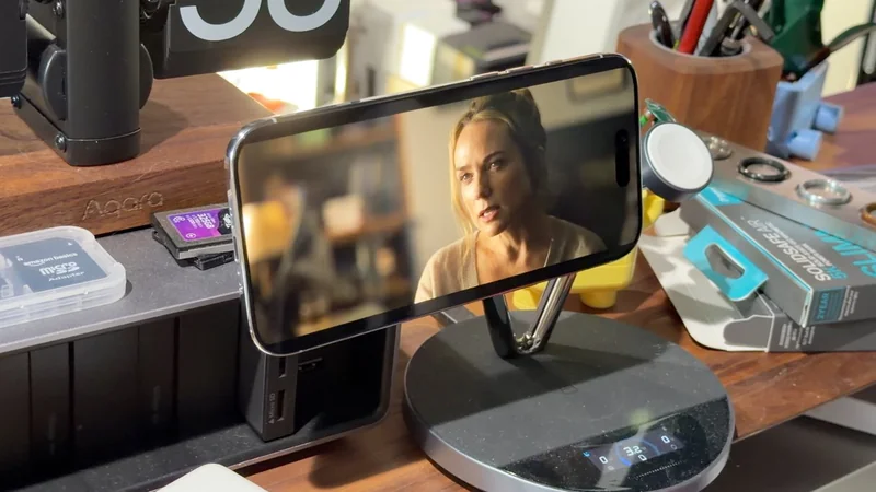 Smartphone on a metal stand playing a video of a woman speaking, surrounded by neatly arranged desk accessories, chargers, and tech gadgets on a wooden workspace