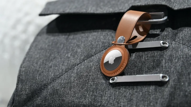 Close-up of an Apple AirTag in a brown leather keychain holder attached to the zipper pull of a gray fabric bag or jacket