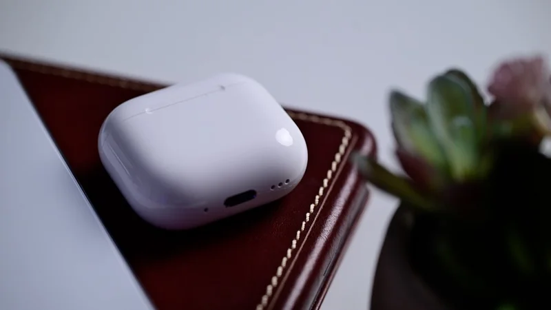 White AirPods 4 charging case resting on a closed brown leather notebook with stitched edges, next to a blurred green succulent plant on a clean, light-colored desk