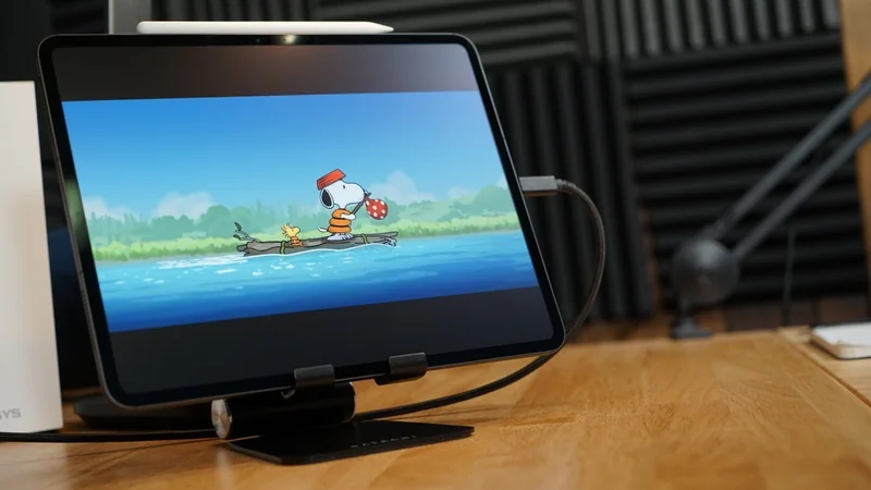 Apple iPad Pro 13-inch on a wooden desk showing Snoopy and Woodstock paddling a canoe on a calm blue river, with studio microphone and soundproof foam panels blurred in the background