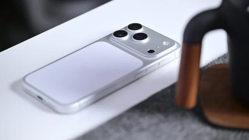 Silver smartphone lying face down on a white surface, showing a large camera module with three lenses, near a dark mug with a wooden handle on a gray textured mat