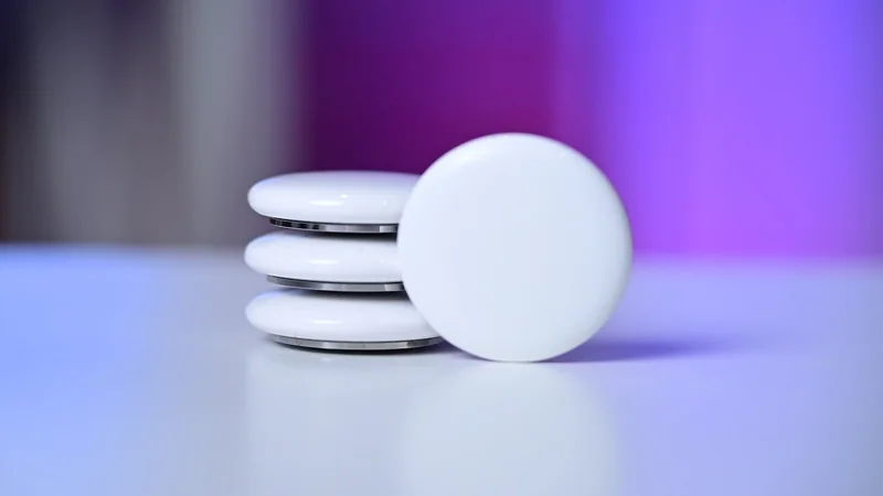 Stack of Apple AirTag trackers on a smooth surface, one standing upright in front, with a blurred purple and blue background