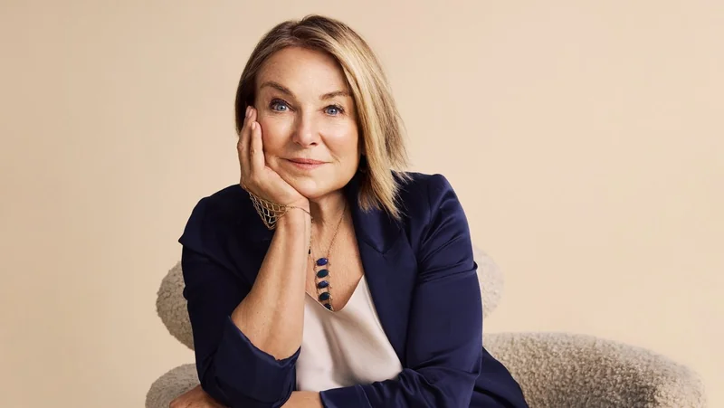 Middle aged woman with blonde bob hair, wearing a navy blazer and white blouse, resting her head on her hand and smiling gently while seated against a plain beige background