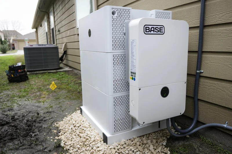 A white-colored residential battery with the name "BASE" on the side sits on the side of a brown-colored house in Houston, Texas.