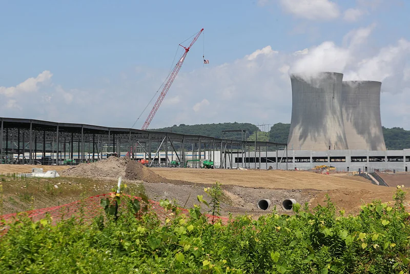 A crane is helping build a large, low building next to the cooling towers of a nuclear power plant.
