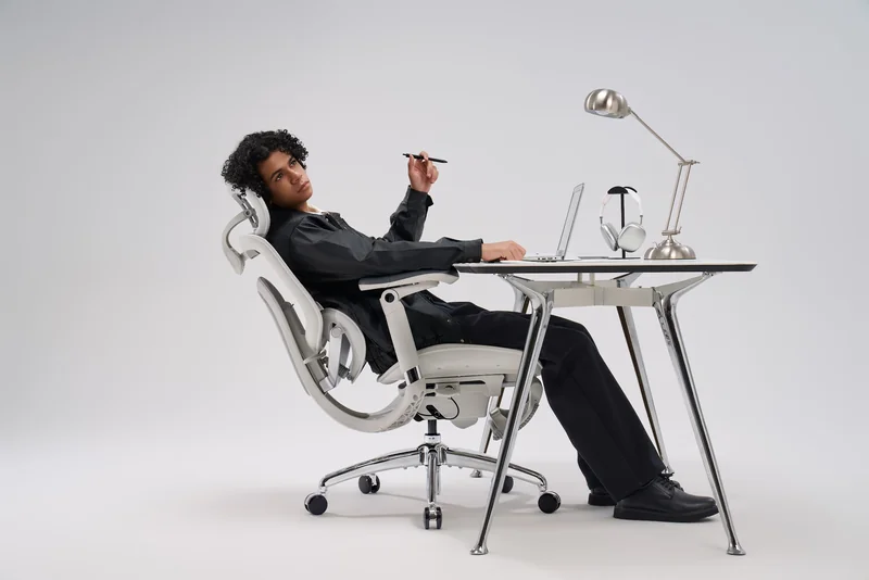 A person reclines in a white sihoo ergonomic chair at a modern desk with a laptop, lamp, and headphones.