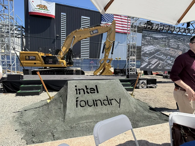 A construction event with a Caterpillar 349 excavator and a mound of dirt displaying 'intel foundry' text, set against flags of California and the United States, with a screen showing an aerial view of a facility.