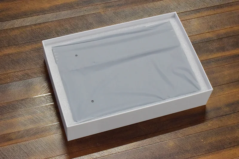 A cardboard box contains an unbranded electronic device wrapped in protective plastic packaging on a wooden floor.