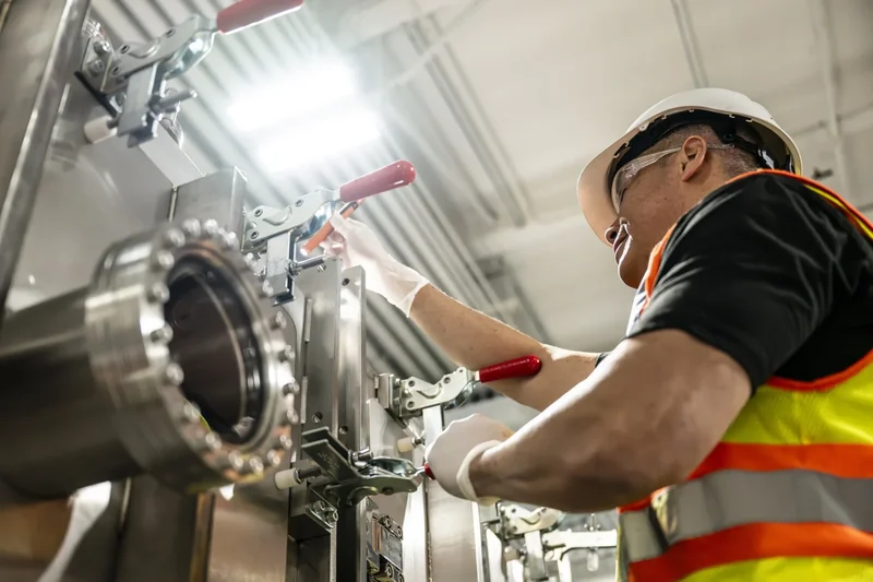A person wearing a hard hat and safety vest operates a large industrial valve in a factory setting.