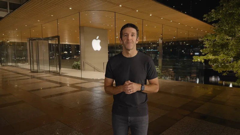 Man standing at night outside a brightly lit modern Apple Store with large glass walls, wooden ceiling, glowing Apple logo, and reflections on the polished plaza floor