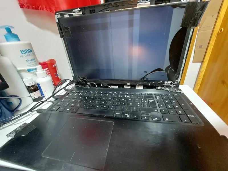 A damaged laptop with a broken screen and exposed internals is positioned on a desk next to various skincare products, including a bottle labeled 'ISDIN Hydration.'