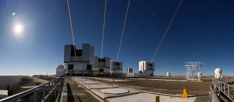 Image of four cylindrical buildings, each with an open slot for the telescope, and each emitting an orange colored laser