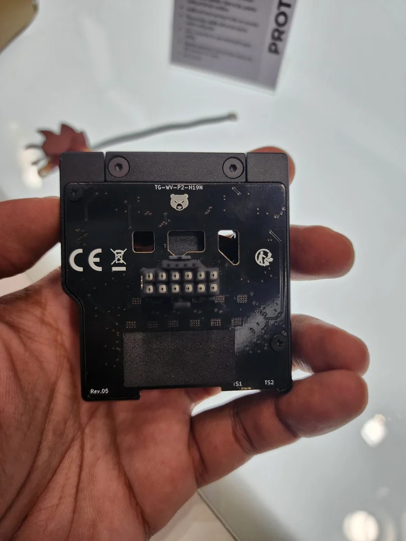 A person holds a component labeled 'TG-WV-P2-H19N' and 'Rev.05', with visible connectors and a partially obscured display stand in the background.
