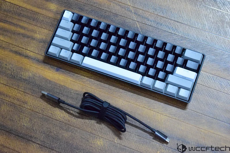 A mechanical keyboard with a gray and black color scheme is placed on a wooden surface alongside a coiled USB cable.