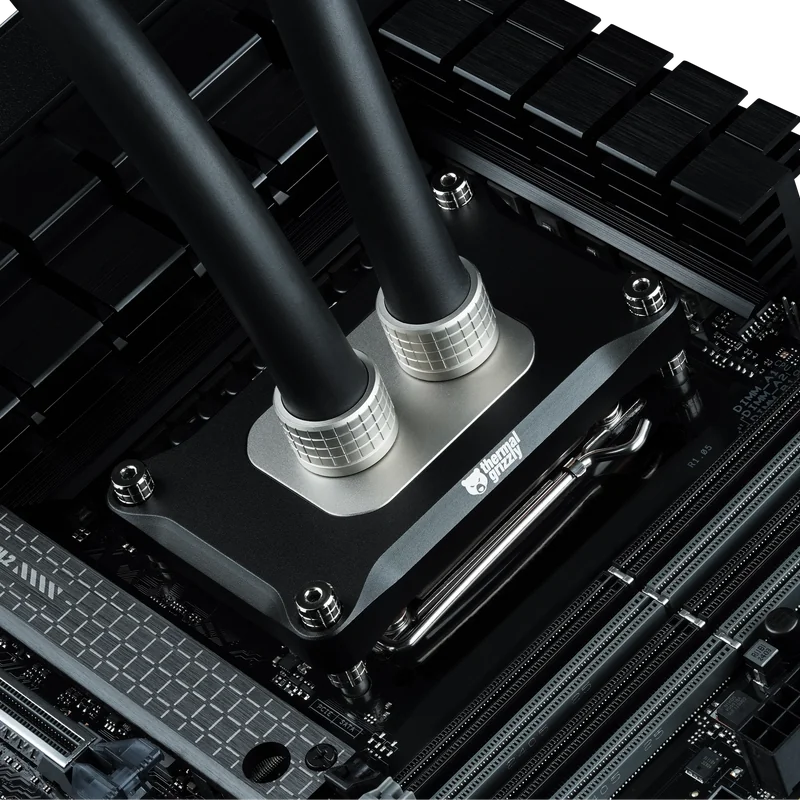 A close-up view of a Thermal Grizzly water block installed on a computer motherboard, showing dual tubes connected to the cooling system.