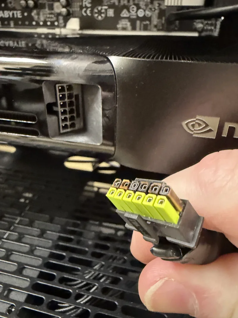 A close-up of a hand holding a yellow and black 12VHPWR connector near a NVIDIA graphics card with a PCIe power port.