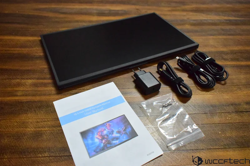 A portable monitor with an '18.5 Inch 1080P Portable Monitor User Manual,' three cables, a power adapter, and a plastic bag with screws placed on a wooden surface.