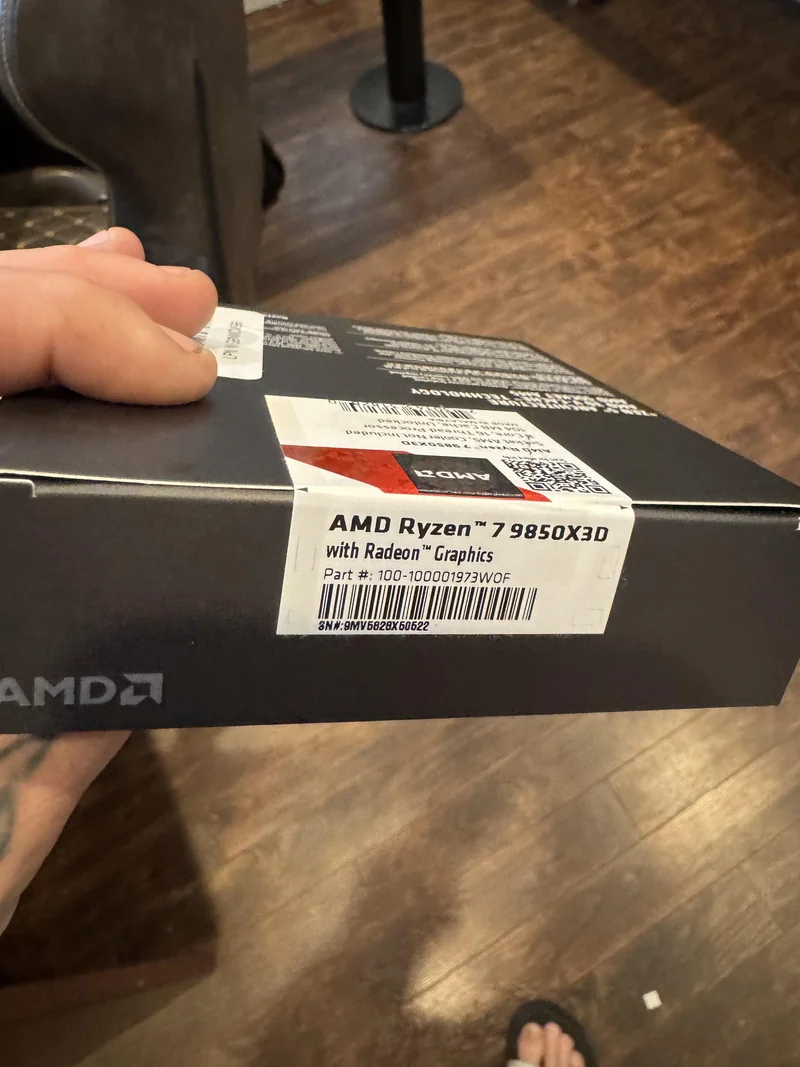 A person holding a box labeled AMD Ryzen 7 9850X3D with Radeon Graphics in a room with hardwood flooring.