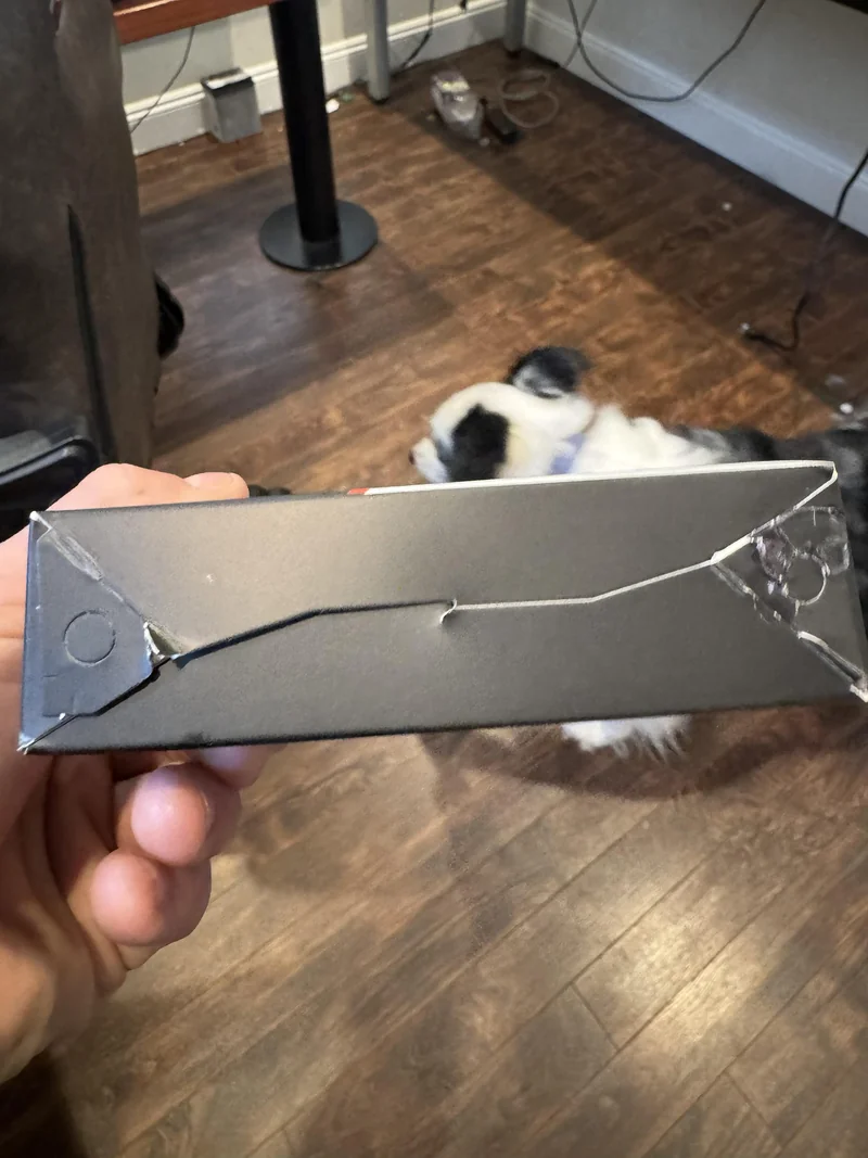 A hand holding a damaged black cardboard box with cracks and tears, while a dog is visible in the background on a wooden floor.