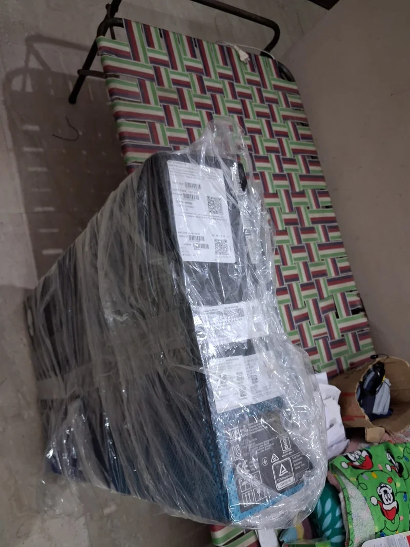 A wrapped, unbranded desktop computer tower sits on a colorful, patterned bed with labels on the packaging showing detailed specifications and QR codes.